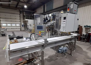 A large industrial packaging machine in a warehouse setting. The conveyor belt and control panel with various buttons and indicators are visible. The background displays additional equipment, ladders, shelves, and large garage doors. The area is well-lit.