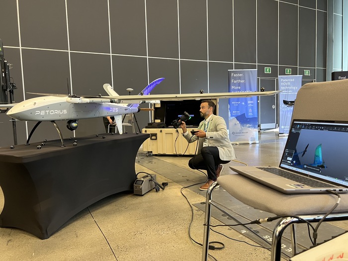 Un homme s'agenouille en examinant une maquette de drone sur une table dans un hall d'exposition. À proximité, un ordinateur portable affiche un rendu de conception du drone. Des bannières d'exposition et des cloisons sont visibles en arrière-plan.